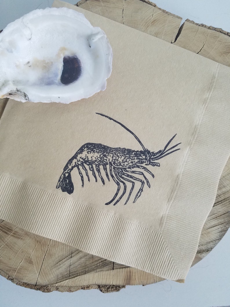 Pode incluir: Um guardanapo de papel bege com uma ilustra&ccedil;&atilde;o de camar&atilde;o preto, colocado em uma fatia de madeira. Uma concha de ostra branca vazia est&aacute; no canto superior esquerdo. O guardanapo tem uma borda canelada.