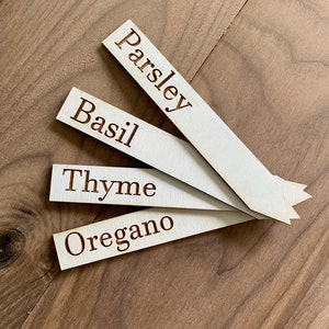 Op de afbeelding: Vier houten tuinmarkeerders met de namen "Peterselie", "Basilicum", "Tijm" en "Oregano" gegraveerd. De lichtgekleurde markeerders liggen op een houten oppervlak.