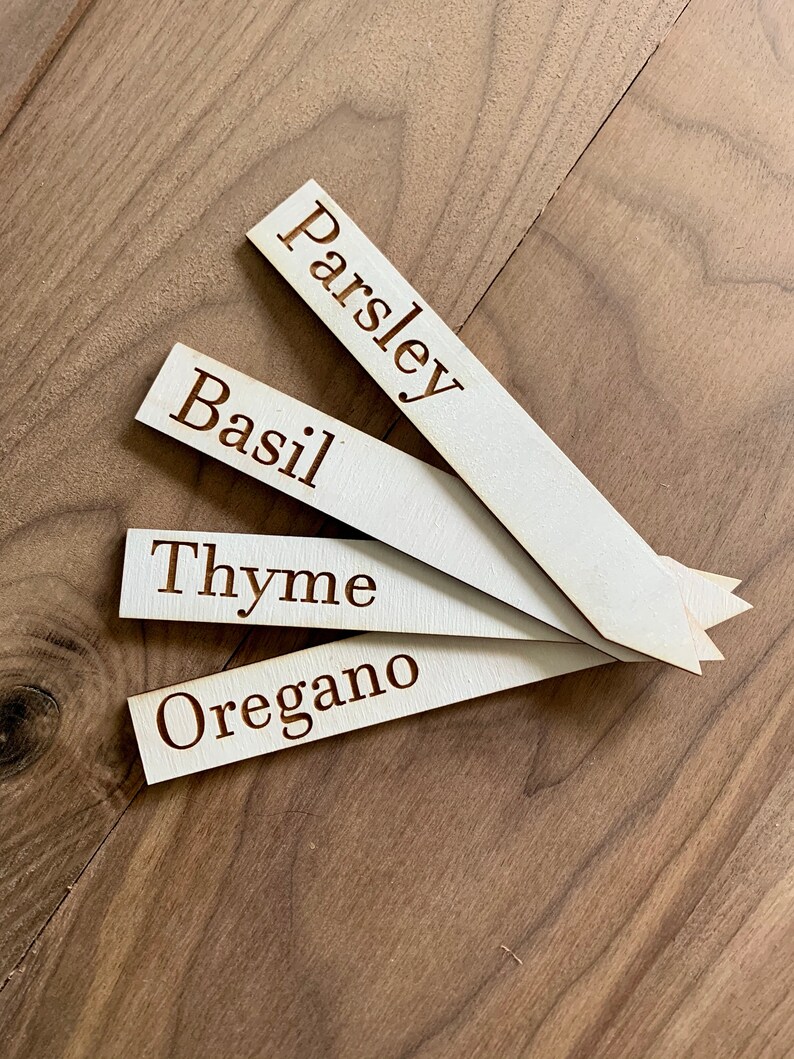 Op de afbeelding: Vier houten tuinmarkeerders met de namen "Peterselie", "Basilicum", "Tijm" en "Oregano" gegraveerd. De lichtgekleurde markeerders liggen op een houten oppervlak.