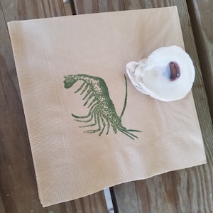 Pode incluir: Uma pilha de guardanapos de papel bege com uma ilustra&ccedil;&atilde;o de lagosta verde e uma concha de ostra branca. Os guardanapos est&atilde;o sobre uma superf&iacute;cie de madeira. O interior da concha de ostra &eacute; castanho escuro.