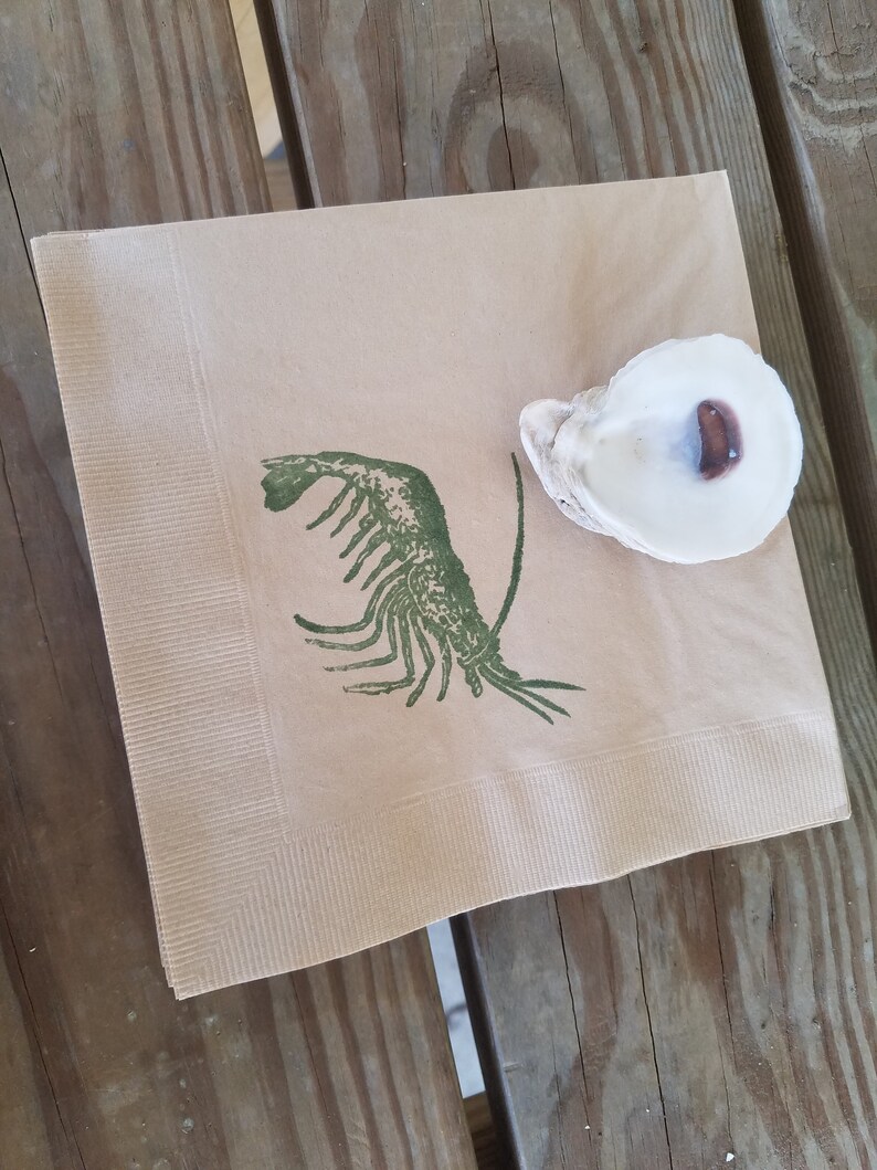 Pode incluir: Uma pilha de guardanapos de papel bege com uma ilustra&ccedil;&atilde;o de lagosta verde e uma concha de ostra branca. Os guardanapos est&atilde;o sobre uma superf&iacute;cie de madeira. O interior da concha de ostra &eacute; castanho escuro.