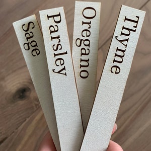 Op de afbeelding: Vier houten tuinlabels met de namen van kruiden: Salie, Peterselie, Oregano en Tijm. De tekst is donkerbruin op een licht houten achtergrond. De labels zijn rechthoekig en licht schuin.