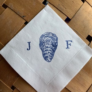 May include: White square paper napkin with a blue illustration of a peanut and the letters "J F". The napkin has a white ribbon border and is set on a brown woven surface.