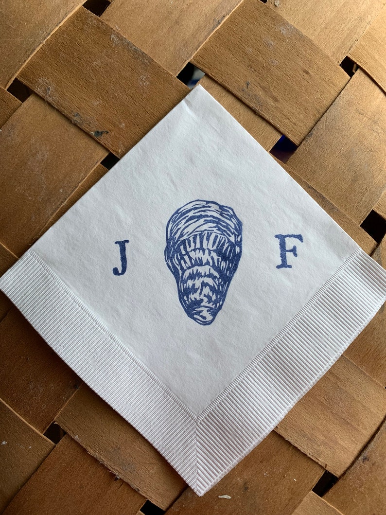 May include: White square paper napkin with a blue illustration of a peanut and the letters "J F". The napkin has a white ribbon border and is set on a brown woven surface.
