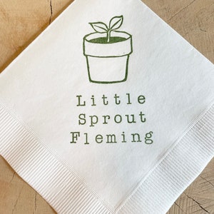 May include: White square napkin featuring a green illustration of a potted plant and the words "Little Sprout Fleming" printed in green. The napkin has a ribbed border and is set on a wooden surface.