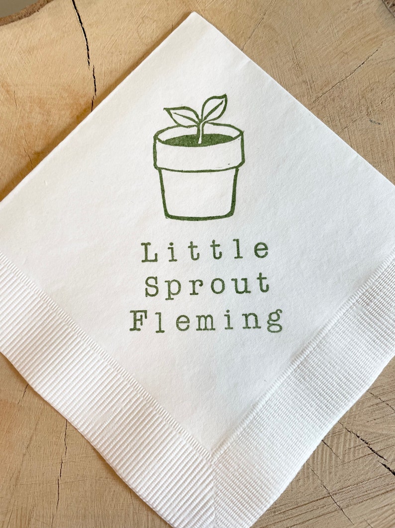 May include: White square napkin featuring a green illustration of a potted plant and the words "Little Sprout Fleming" printed in green. The napkin has a ribbed border and is set on a wooden surface.