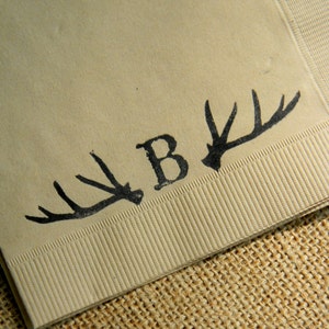 May include: Beige paper napkin with a black antler design and the letter "B". The napkin has a textured surface and is folded. The design is printed in black ink. The napkin is on a burlap surface.