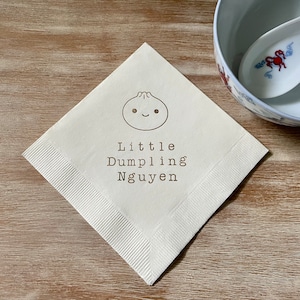 May include: A square, off-white napkin with a smiling dumpling illustration and the text "Little Dumpling Nguyen" in brown. A white bowl and spoon with a red and blue design are in the background.