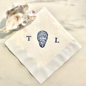 May include: White linen napkin with a blue oyster illustration and the letters "T" and "L". The napkin has a white ribbon border and is next to a natural oyster shell. A coastal themed item.