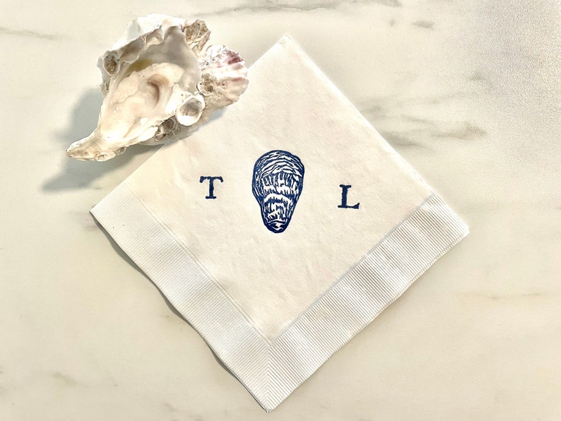 May include: White linen napkin with a blue oyster illustration and the letters "T" and "L". The napkin has a white ribbon border and is next to a natural oyster shell. A coastal themed item.