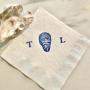 May include: White paper napkin with a blue oyster illustration and the letters "T" and "L". The napkin has a ribbed border and is next to an oyster shell.