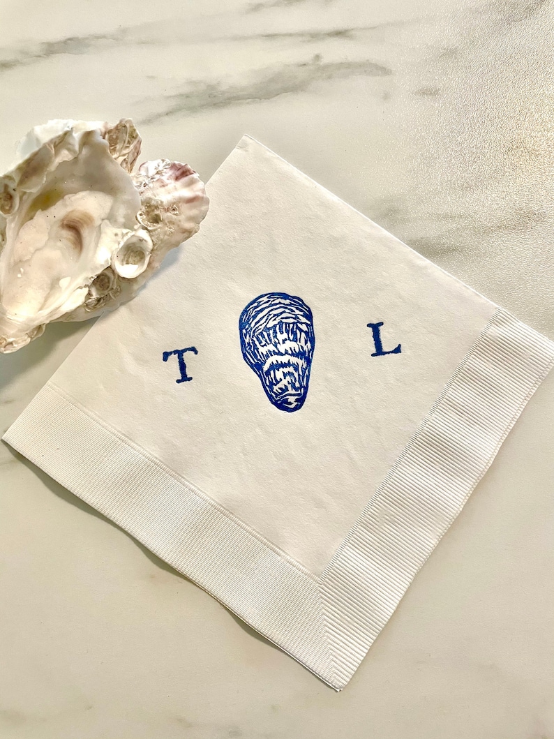 May include: White paper napkin with a blue oyster illustration and the letters "T" and "L". The napkin has a ribbed border and is next to an oyster shell.