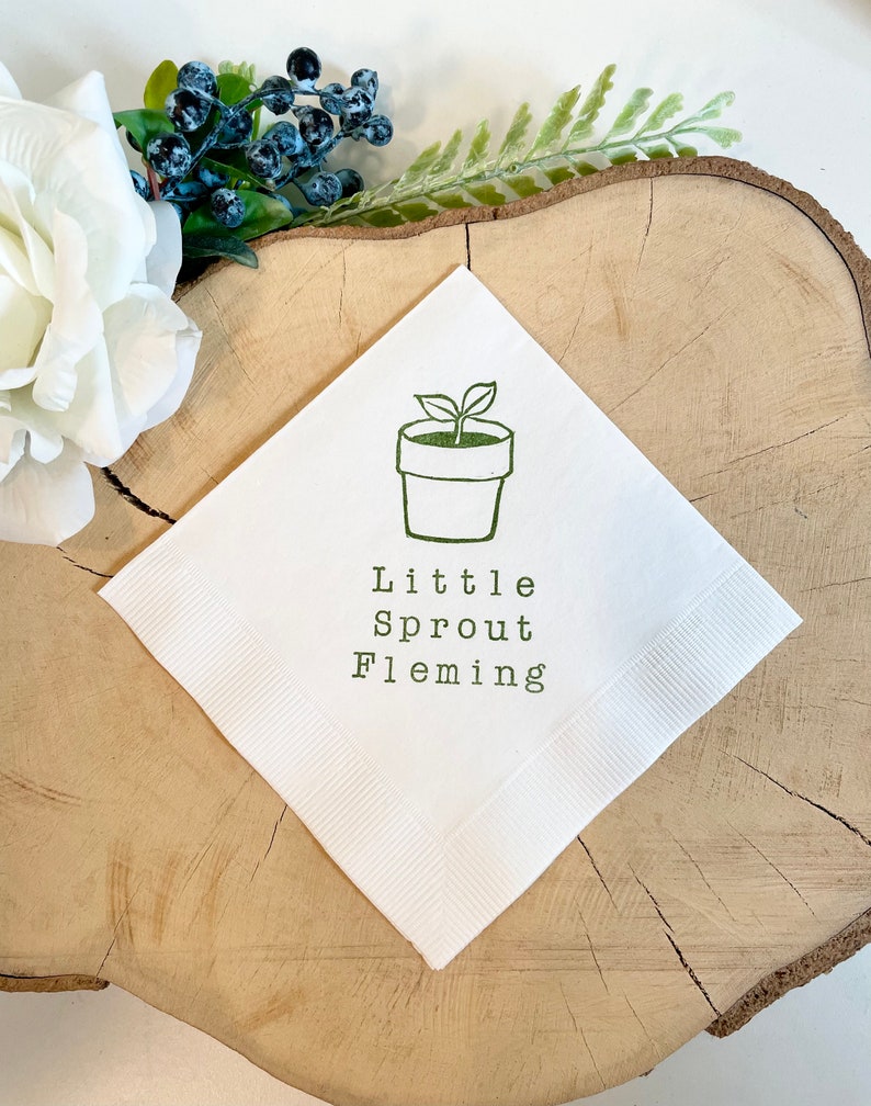 May include: White square napkin featuring a green illustration of a potted plant and the text "Little Sprout Fleming." The napkin has a ribbed border. It is displayed on a wooden surface with floral decorations.