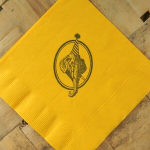 May include: A yellow paper napkin with a black illustration of an elephant wearing a party hat inside a circle. The napkin is square and has a textured surface.