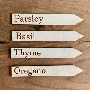 Op de afbeelding: Vier houten plantenlabels met puntige uiteinden, elk gemarkeerd met een andere kruidnaam in bruine letters: Peterselie, Basilicum, Tijm en Oregano. De labels zijn gerangschikt op een houten oppervlak.