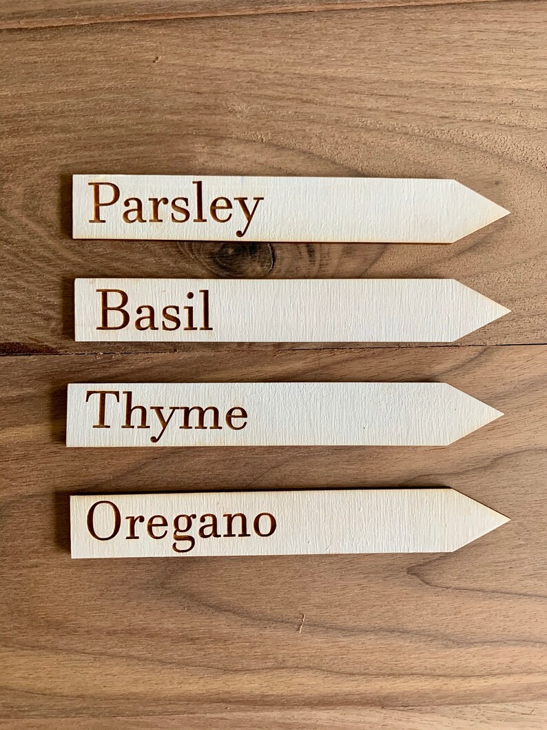 Op de afbeelding: Vier houten plantenlabels met puntige uiteinden, elk gemarkeerd met een andere kruidnaam in bruine letters: Peterselie, Basilicum, Tijm en Oregano. De labels zijn gerangschikt op een houten oppervlak.