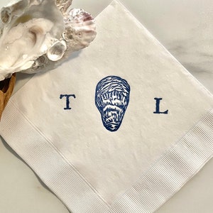 May include: White linen napkin with a navy blue oyster illustration and the letters "T" and "L". The napkin has a ribbed border and is folded diagonally. An oyster shell and driftwood are in the background.