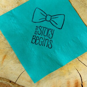 Op de afbeelding: Een turquoise servet met een zwarte vlinderdas illustratie en de tekst "and the story begins". Het servet ligt op een houten oppervlak met een jute rand. De vlinderdas staat boven de tekst.
