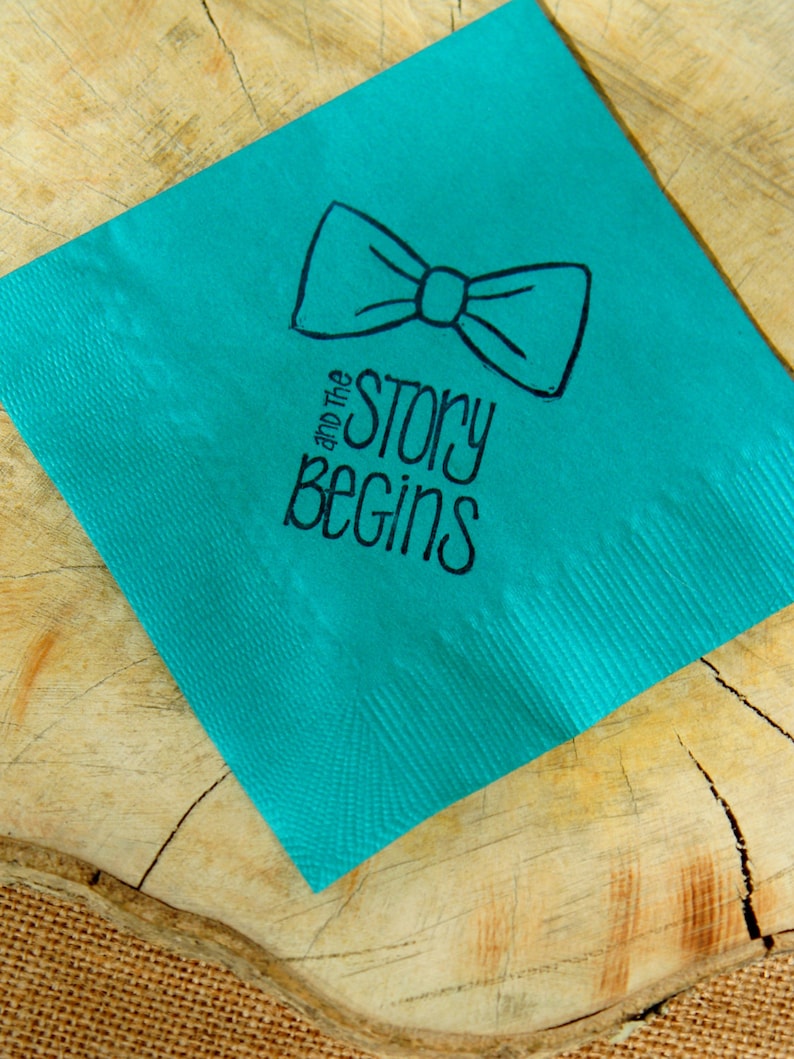 Op de afbeelding: Een turquoise servet met een zwarte vlinderdas illustratie en de tekst "and the story begins". Het servet ligt op een houten oppervlak met een jute rand. De vlinderdas staat boven de tekst.