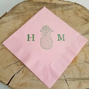 May include: A pink square napkin with a green pineapple illustration and the letters "H" and "M". The napkin has a ribbed border and is displayed on a wooden surface. The pineapple design is centered.