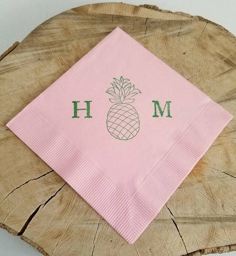May include: A pink square napkin with a green pineapple illustration and the letters "H" and "M". The napkin has a ribbed border and is displayed on a wooden surface. The pineapple design is centered.