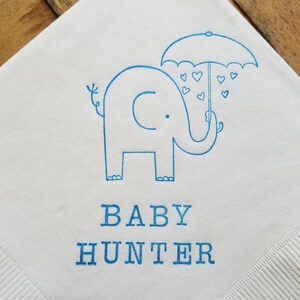 May include: White paper napkin with a blue illustration of an elephant holding an umbrella with heart-shaped raindrops. The words "BABY HUNTER" are printed in blue below the elephant.