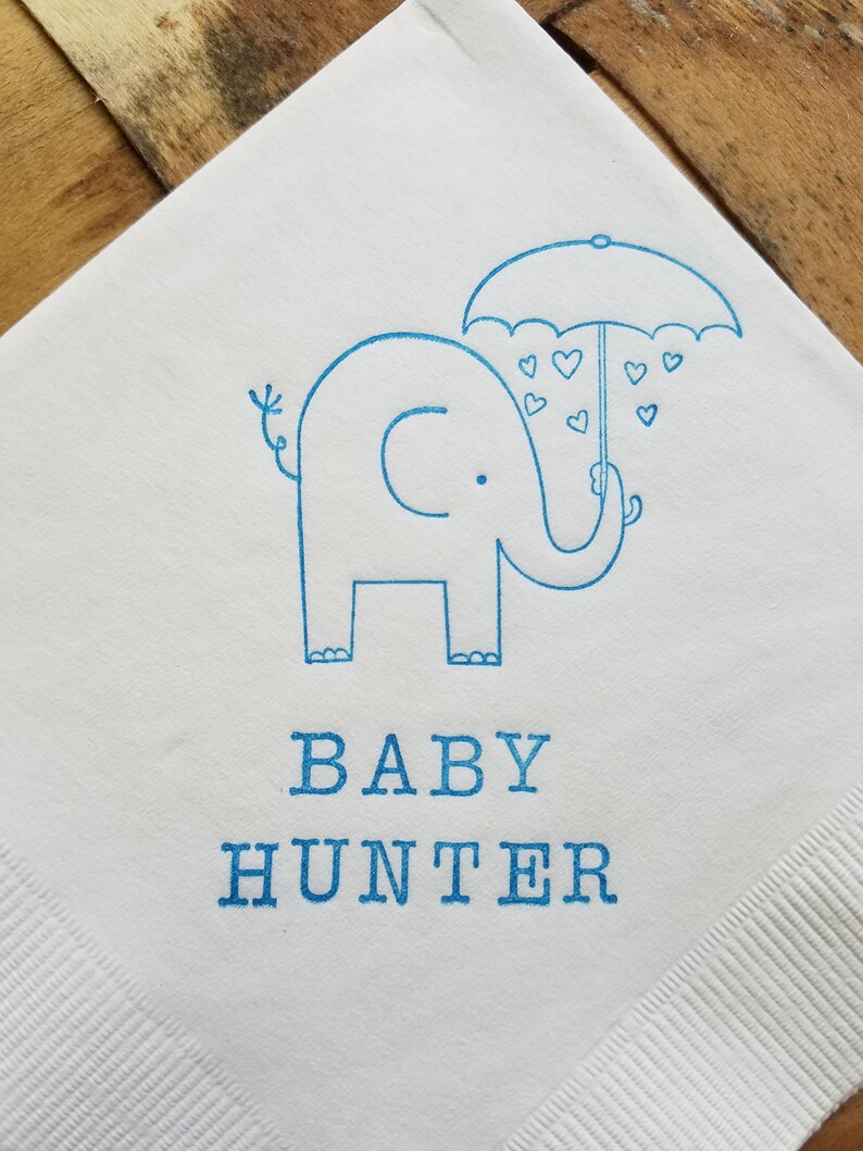 May include: White paper napkin with a blue illustration of an elephant holding an umbrella with heart-shaped raindrops. The words "BABY HUNTER" are printed in blue below the elephant.