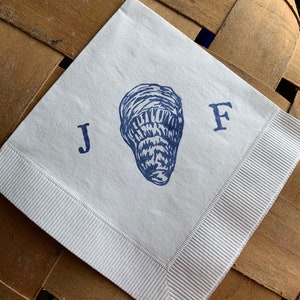 May include: White square paper napkin with a blue oyster illustration and the letters "J" and "F". The napkin has a white ribbon border and is set against a woven wooden background.