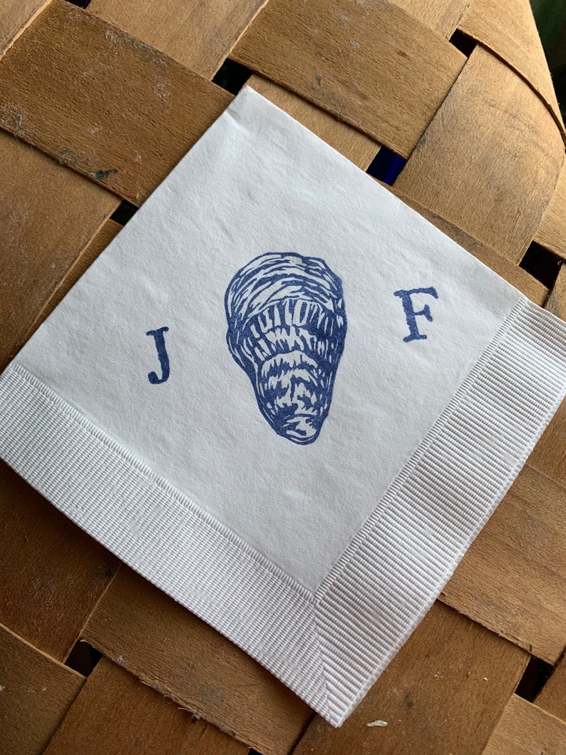 May include: White square paper napkin with a blue oyster illustration and the letters "J" and "F". The napkin has a white ribbon border and is set against a woven wooden background.