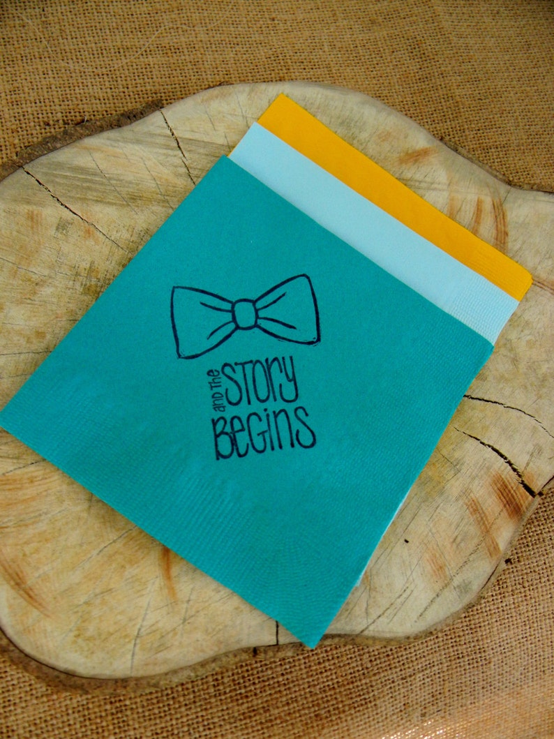 Op de afbeelding: Een turquoise notitieboek met een vlinderdas-illustratie en de woorden "and the story begins" in het zwart. Het notitieboek heeft lichtblauwe en gele pagina's. Het notitieboek ligt op een houten oppervlak.