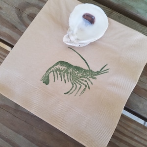 Pode incluir: Uma guardanapo de papel quadrado, bege, com uma ilustra&ccedil;&atilde;o de camar&atilde;o verde e uma concha de ostra branca com interior castanho. O guardanapo est&aacute; sobre uma superf&iacute;cie de madeira.