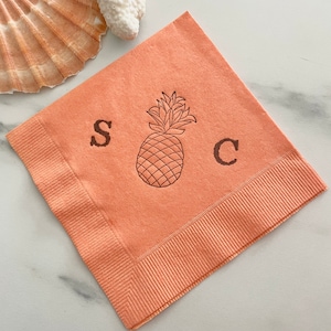 May include: Coral-colored cocktail napkin with a pineapple illustration and the letters "S" and "C". The napkin has a ribbed border and is displayed with seashells. Ideal for a tropical-themed event.