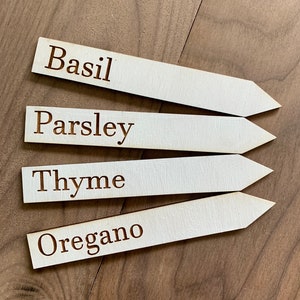 Op de afbeelding: Vier houten tuinmarkeringen met puntige uiteinden, met de labels "Basilicum", "Peterselie", "Tijm" en "Oregano" in een donkerbruin lettertype. De markeringen zijn lichtbeige en liggen op een donkerbruin houten oppervlak. Voor kruidentuinen.