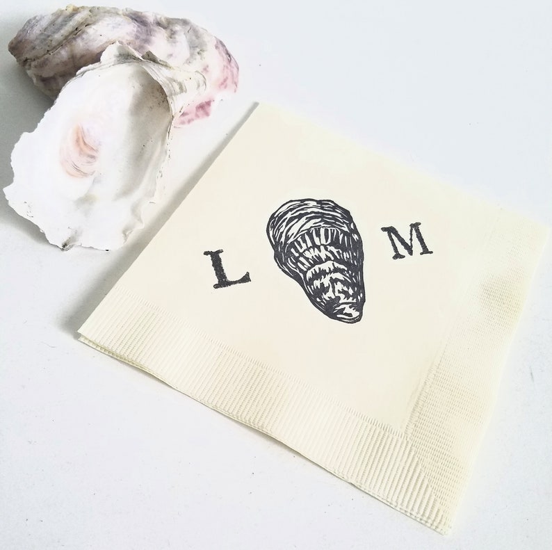 May include: Off-white paper napkin with a black oyster illustration and the letters "L" and "M". Two oyster shells are in the background. The napkin is folded and has a textured border.