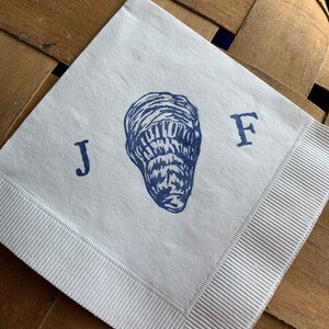 May include: Square white paper napkin with a blue illustration of an oyster and the letters "J" and "F". The napkin has a white ribbon border. The napkin is on a brown woven surface.