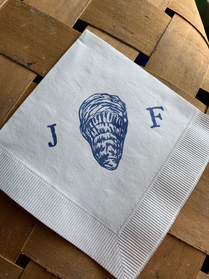 May include: Square white paper napkin with a blue illustration of an oyster and the letters "J" and "F". The napkin has a white ribbon border. The napkin is on a brown woven surface.