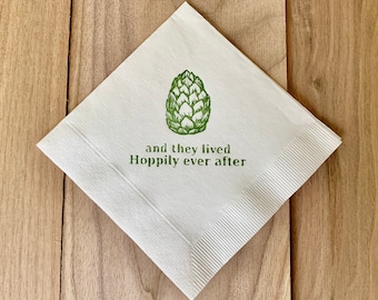 May include: A white square napkin with a green hop illustration and the phrase "and they lived Hoppily ever after." The napkin has a textured border and is set on a wooden surface.