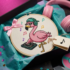 Könnte beinhalten: Ein runder Stickrahmen aus Holz mit einem Kreuzstichmuster eines rosafarbenen Flamingos, der eine türkisfarbene Kappe und Kopfhörer trägt und an einem DJ-Plattenspieler steht. Eine hellrosa Satinschleife ist am Rahmen befestigt, dazu ein rosa Kopfhörer.