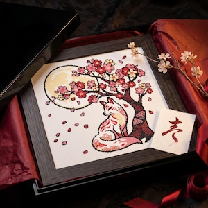 May include: Cross-stitch art of a fox under a cherry blossom tree, framed in a dark wood. The artwork features a full moon and falling petals. The piece is presented in a black box with red tissue paper.