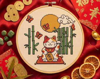 May include: A circular wooden embroidery hoop containing a cross-stitch design of a calico beckoning cat sitting on a red mat, surrounded by green bamboo stalks, orange butterflies, a yellow full moon, and a white cloud on a cream fabric background.