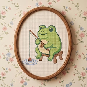 Puede incluir: Arte de punto de cruz de una rana verde pescando, enmarcado en un aro ovalado de madera. La rana está sentada en un pequeño taburete, sosteniendo una caña de pescar. El fondo es un papel pintado floral.