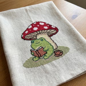 May include: A beige linen tea towel with a cross-stitched design. The design features a green frog reading a book under a red mushroom with white spots. A small teacup is next to the frog. The towel is folded on a wooden surface.