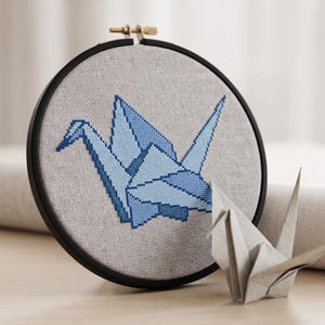 May include: A black embroidery hoop containing light gray fabric with a cross-stitched design of a blue origami crane. The crane is rendered in various shades of blue thread. A small, folded paper origami crane sits in front of the hoop.