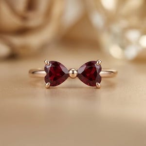 May include: A gold ring featuring two deep red, heart-shaped gemstones arranged to form a bow shape. The stones are set in a polished gold band, with a small gold sphere positioned at the center between the two heart-shaped gems.