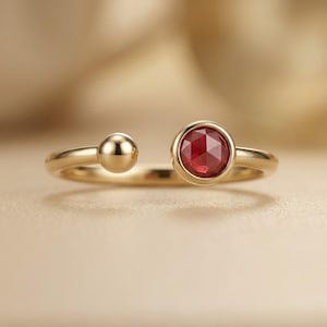 May include: A gold open ring featuring a round, faceted red gemstone set in a bezel on one end and a smooth, polished gold sphere on the other. The band is thin and circular, showcasing a minimalist design with a warm, metallic yellow finish.