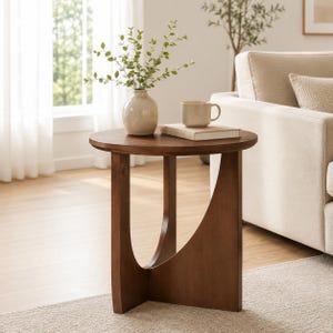 May include: A round wooden side table with a dark brown finish, featuring a unique curved base design. On the tabletop sits a light-colored ceramic vase filled with green leafy branches and a beige ceramic mug resting on a closed book.