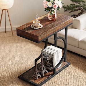 May include: A C-shaped side table featuring a dark brown wood grain top and base with a black metal frame. The table includes a lower shelf holding several books and two small Eiffel Tower figurines, while the top surface holds a coffee cup.