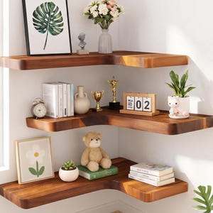 May include: Three L-shaped floating corner shelves made of dark wood with rounded edges. The shelves hold various decorative items including a framed leaf print, a vase with flowers, books, a clock, trophies, a calendar showing 02, a teddy bear, and plants.
