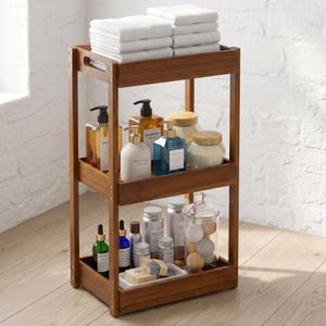 May include: A three-tiered wooden bathroom storage cart with a warm brown finish. The top shelf holds a stack of folded white towels. The middle shelf contains various shampoo and lotion bottles. The bottom shelf holds small jars and soap.