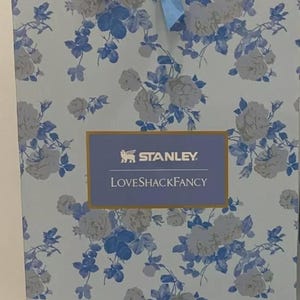 May include: A box with a floral pattern in light blue and white, tied with a blue bow. The box displays the "STANLEY" and "LOVESHACKFANCY" logos. The floral design features white roses and blue leaves.